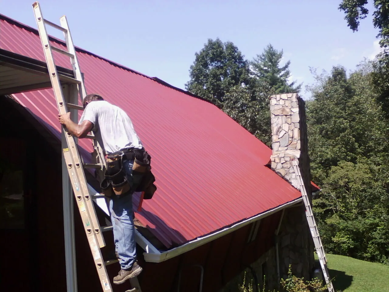 Roofer on a ladder beside a red metal roof and stone chimney — Soffit & Fascia Repair by Roofing Brothers in Harrisville