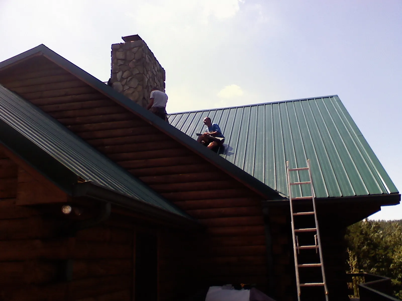 Roofers installing green metal panels beside a stone chimney on a log home in Harrisville